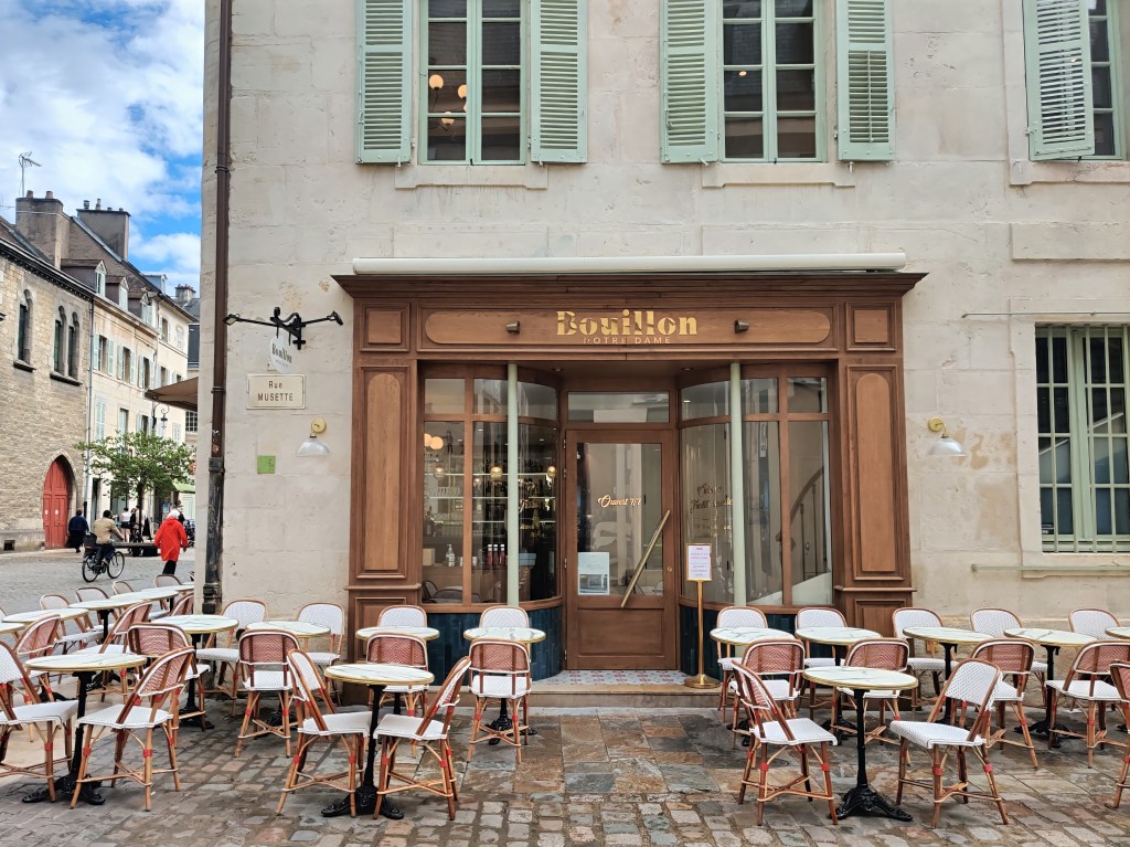 Beautiful cafe facade in Dijon, France with green shutters and red and white chairs. 