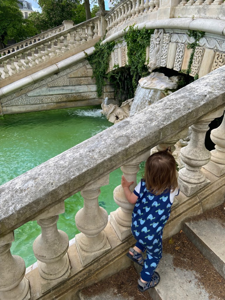 Toddler in turtle overalls looking at the fountain at Jardin Darcy in Dijon, France. 