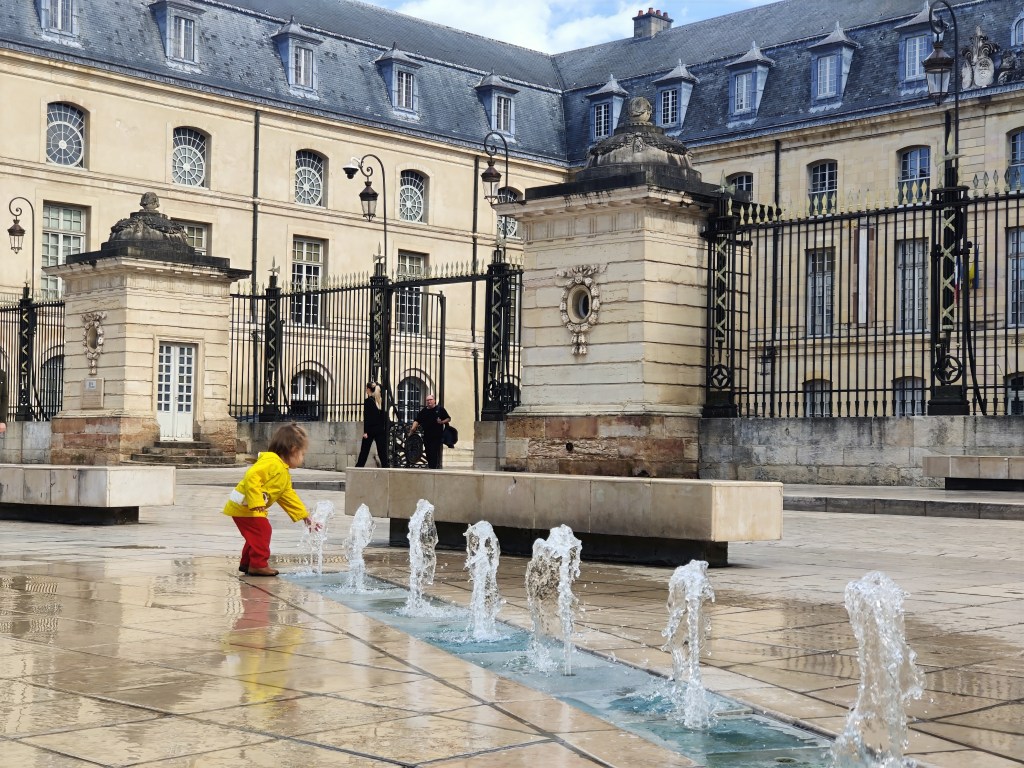 Toddler in a yellow rain jacket putting his hand into the fountains at Place de la Liberation in Dijon, France