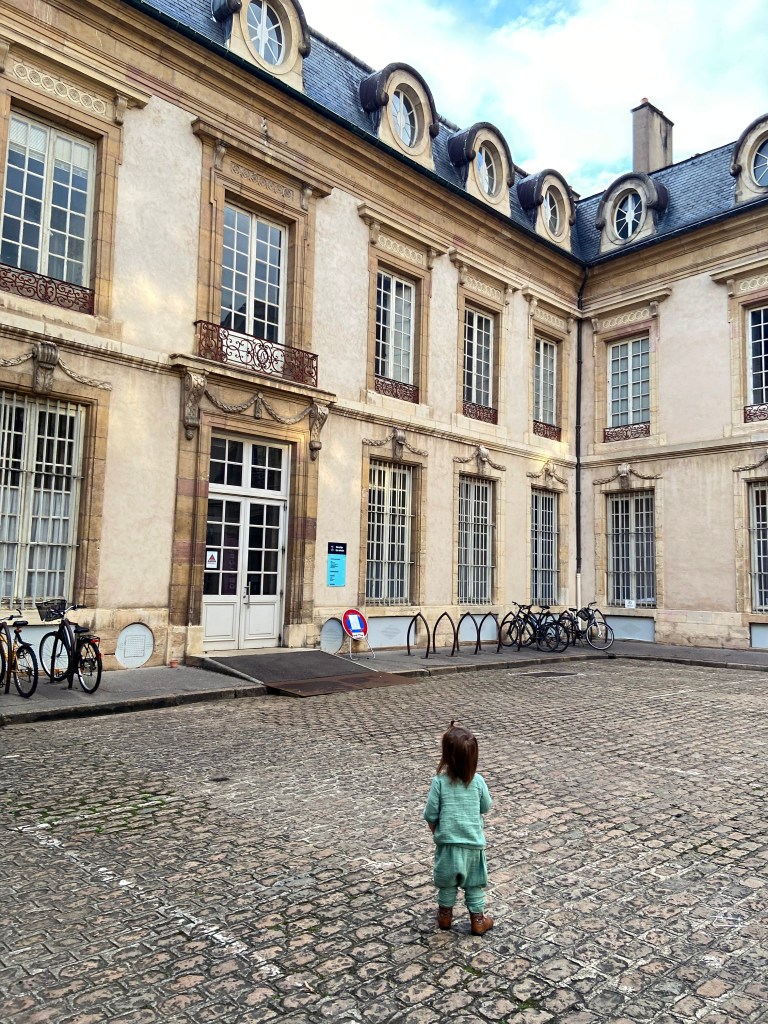 Toddler seen from behind standing in a square in Dijon, France. They're wearing a sage green linen outfit and cowboy boots.