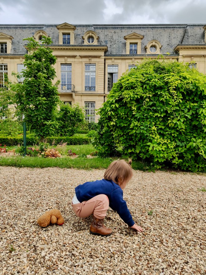Toddler playing in the stones at the Square Albert Schweitzer in Paris. Teddy bear next to them and an opulent hotel in view.